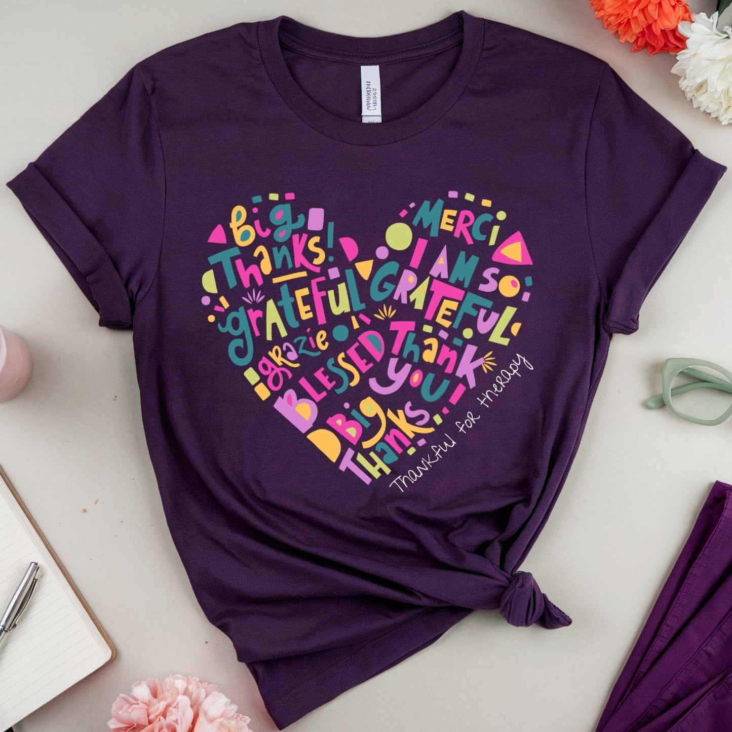 Purple t-shirt with colorful heart-shaped text design on a light surface with flowers and sunglasses.
