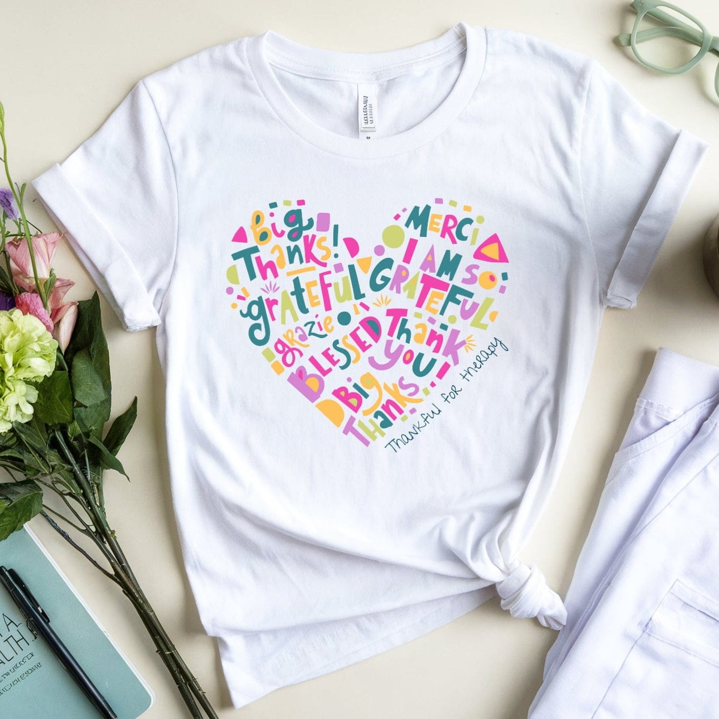 White t-shirt with colorful text design on a table with flowers, coffee, and glasses.