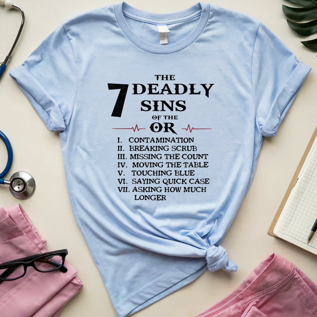 A light blue graphic T-shirt laid flat on a beige surface, surrounded by medical-themed props including pink scrubs, a stethoscope, glasses, a notebook, and a green leaf. The shirt has black printed text reading 'THE 7 DEADLY SINS OF THE OR' with a list below.