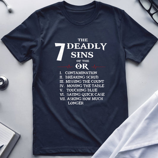 A navy blue graphic t-shirt laid flat on a light gray surface, surrounded by medical-themed props including a stethoscope, glasses, a notebook, and a white lab coat. The shirt features a white printed graphic listing 'The 7 Deadly Sins of the OR'.