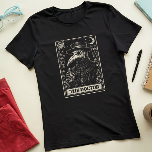 A black short-sleeve crewneck t-shirt featuring a vintage-style tarot card graphic of 'The Doctor' in a plague doctor mask holding a glass, styled in a flat lay with red shorts, light blue glasses, a notebook, and a coffee cup on a cream background.