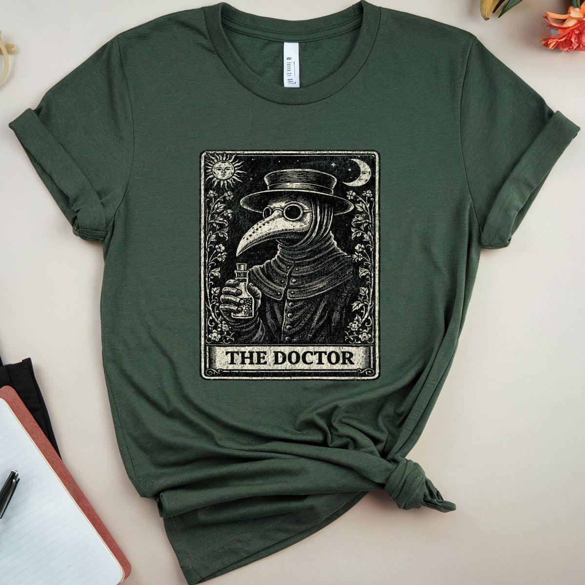 An olive green short-sleeve t-shirt with a black and white tarot card graphic of a plague doctor labeled 'THE DOCTOR', styled in a flat-lay arrangement with a notebook, glasses, and flowers on a light beige background.