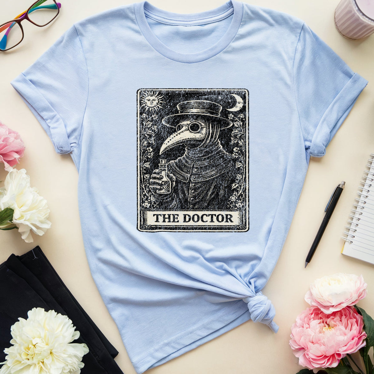 A light blue graphic T-shirt laid flat on a beige surface, styled with flowers, glasses, a notebook, a pen, and a drink. The shirt has a large black rectangular print featuring a plague doctor illustration and the words 'THE DOCTOR'.