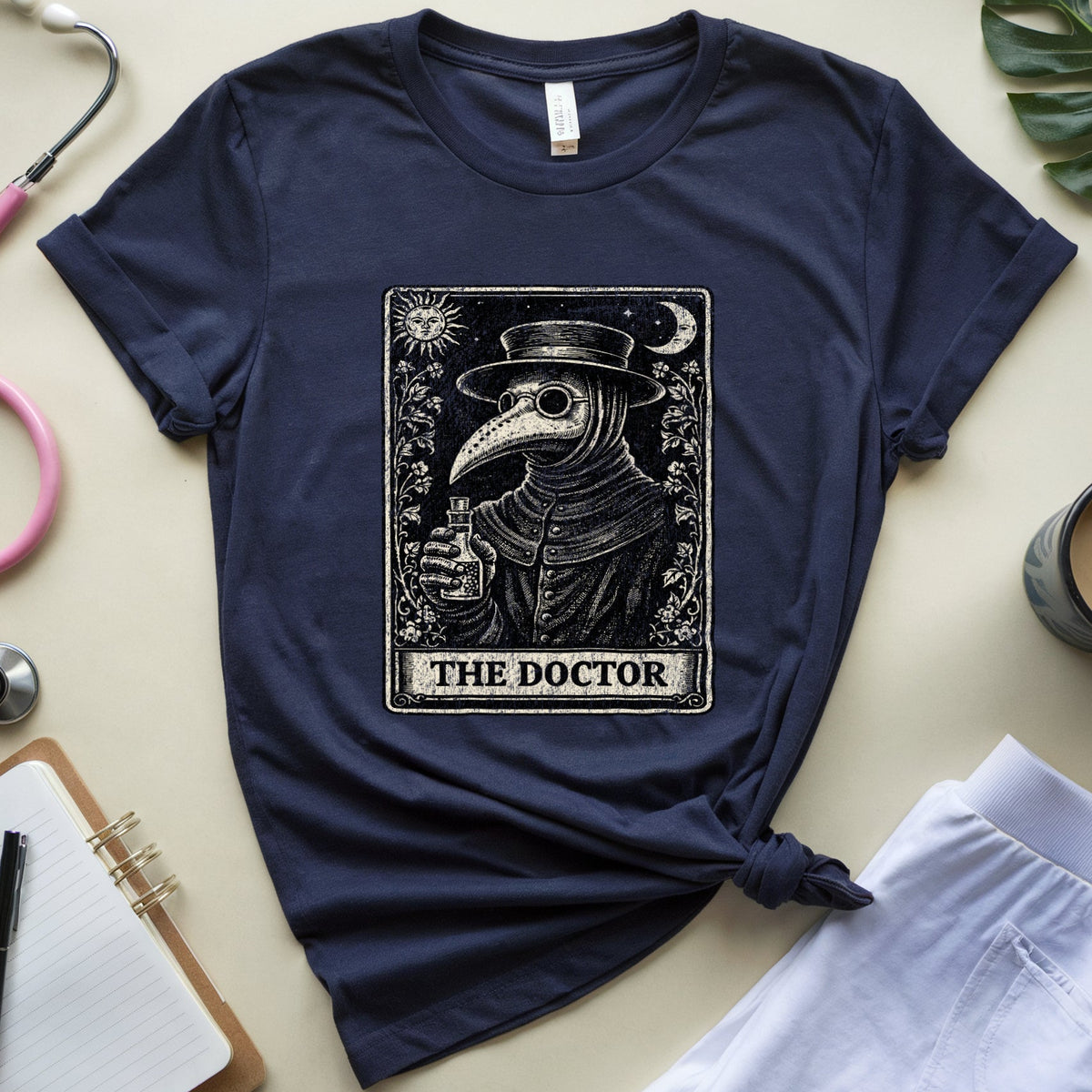 A navy blue short-sleeve T-shirt with a vintage-style tarot card graphic of a plague doctor labeled 'THE DOCTOR,' styled in a flat lay with a pink stethoscope, notebook, plant leaf, coffee cup, and white shorts on a light beige background.