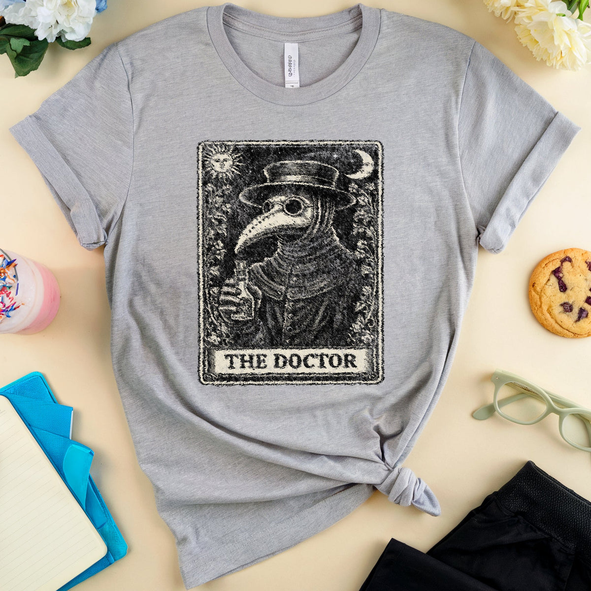 A heather gray short-sleeve T-shirt laid flat on a light beige surface, styled with flowers, a drink, notebook, glasses, and a cookie around it. The shirt has a large black tarot-style graphic of a bird doctor holding a flask, with the text 'THE DOCTOR' on the front.