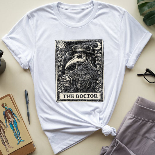 A white short-sleeve T-shirt laid flat on a light beige surface, styled with a black-and-white tarot-style graphic reading 'THE DOCTOR' on the front. Surrounding props include a coffee mug, notebook, pen, eyeglasses, folded gray shorts, and a green leaf.