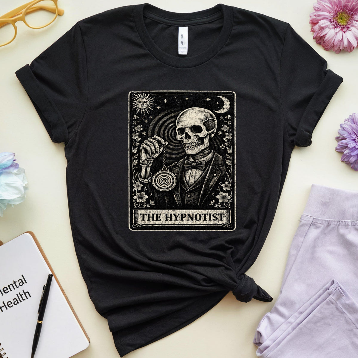 A black graphic t-shirt featuring a tarot card design of 'The Hypnotist' with a skeleton, styled in a flat-lay arrangement with flowers, glasses, a notebook, and sweatpants on a light beige background.