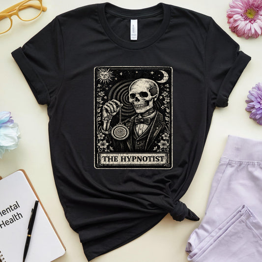 A black graphic t-shirt featuring a tarot card design of 'The Hypnotist' with a skeleton, styled in a flat-lay arrangement with flowers, glasses, a notebook, and sweatpants on a light beige background.