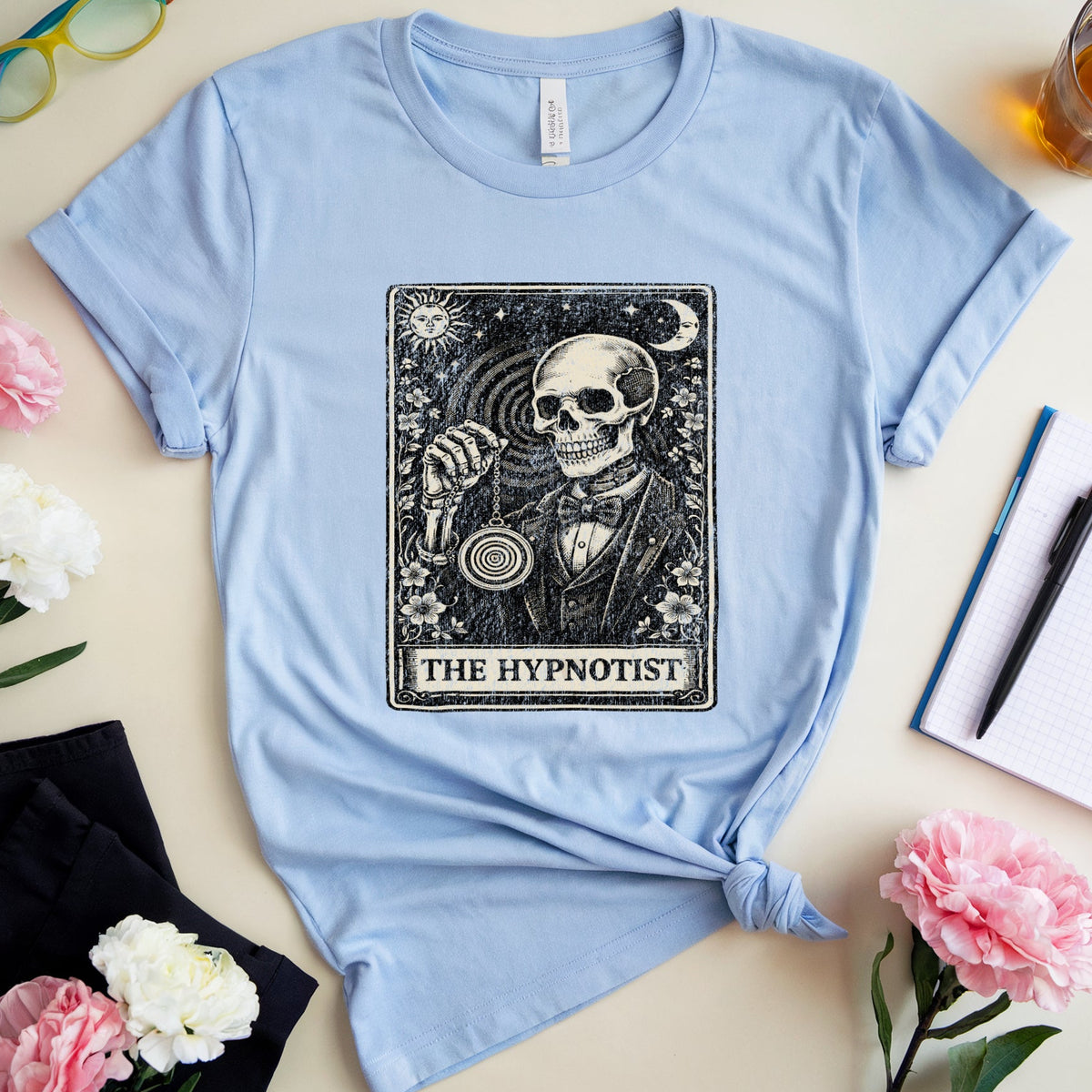 A light blue graphic T-shirt laid flat on a cream surface, styled with flowers, glasses, a drink, notebook, and pen. The shirt has a large black tarot-style illustration reading 'THE HYPNOTIST' on the front.