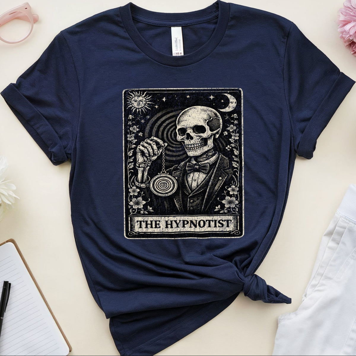A navy blue graphic T-shirt laid flat on a light beige surface, styled with pink flowers, pink glasses, a notebook with a pen, a folded white garment, and a mug of coffee. The shirt has a large black-and-white tarot-style print reading “THE HYPNOTIST” with a skull and circular device graphic.