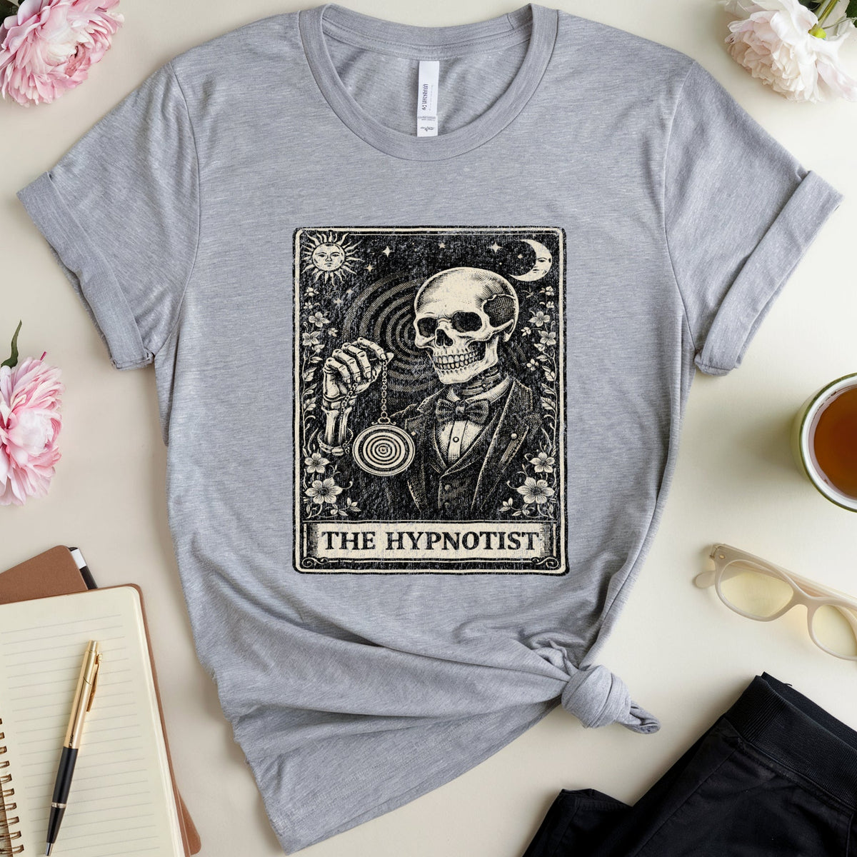 A heather gray short-sleeve t-shirt with a black and white tarot card graphic reading 'THE HYPNOTIST' featuring a skull and moon, styled in a flat lay with pink flowers, a notebook, glasses, and a cup of tea on a light beige background.