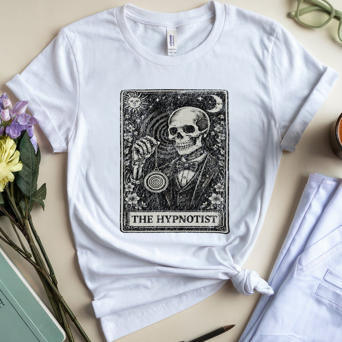A white graphic T-shirt laid flat on a beige surface, styled with flowers, folded light blue pants, a cup of latte, glasses, a pen, and a mint notebook. The shirt features a large black tarot-style illustration reading 'THE HYPNOTIST' with a skull and ornate border.