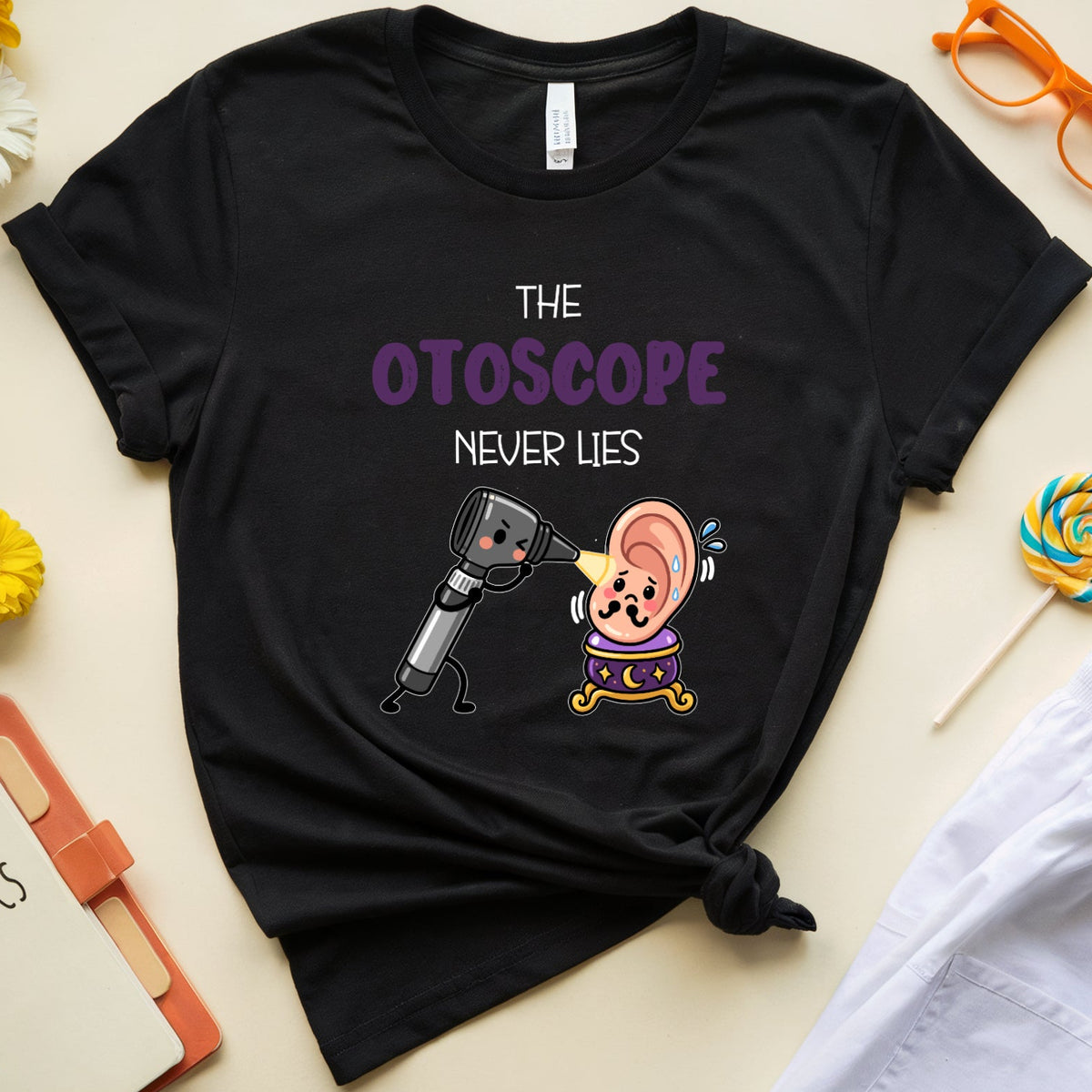 A black graphic t-shirt featuring a cartoon otoscope and ear with the text 'THE OTOSCOPE NEVER LIES' in purple and white. The shirt is styled in a flat lay on a light beige background with medical-themed props including a notebook, white pants, orange glasses, and flowers.