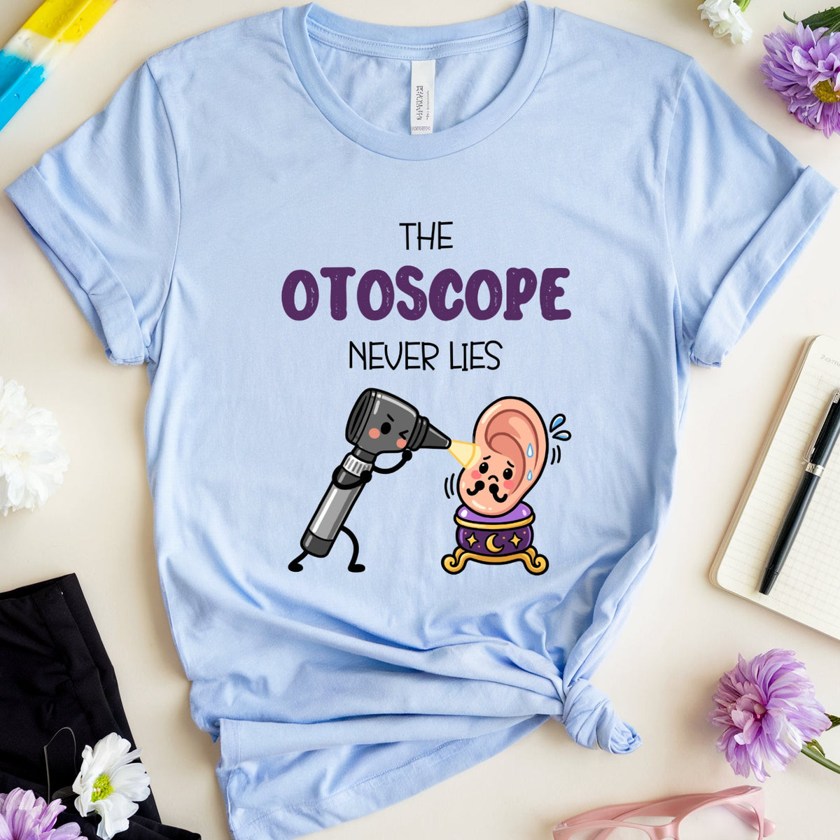 A light blue graphic t-shirt laid flat on a beige surface, surrounded by decorative props including flowers, a popsicle, glasses, shorts, and a notebook. The shirt features a cartoon ear being examined by an otoscope with the text 'THE OTOSCOPE NEVER LIES'.