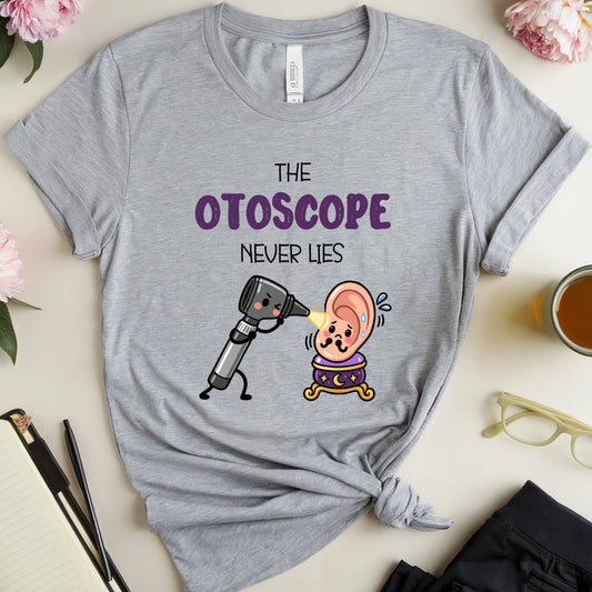 A heather gray graphic T-shirt laid flat on a light beige surface with flowers, a notebook, glasses, a mug, and folded pants around it. The shirt has a cartoon otoscope and ear design with the text 'THE OTOSCOPE NEVER LIES.