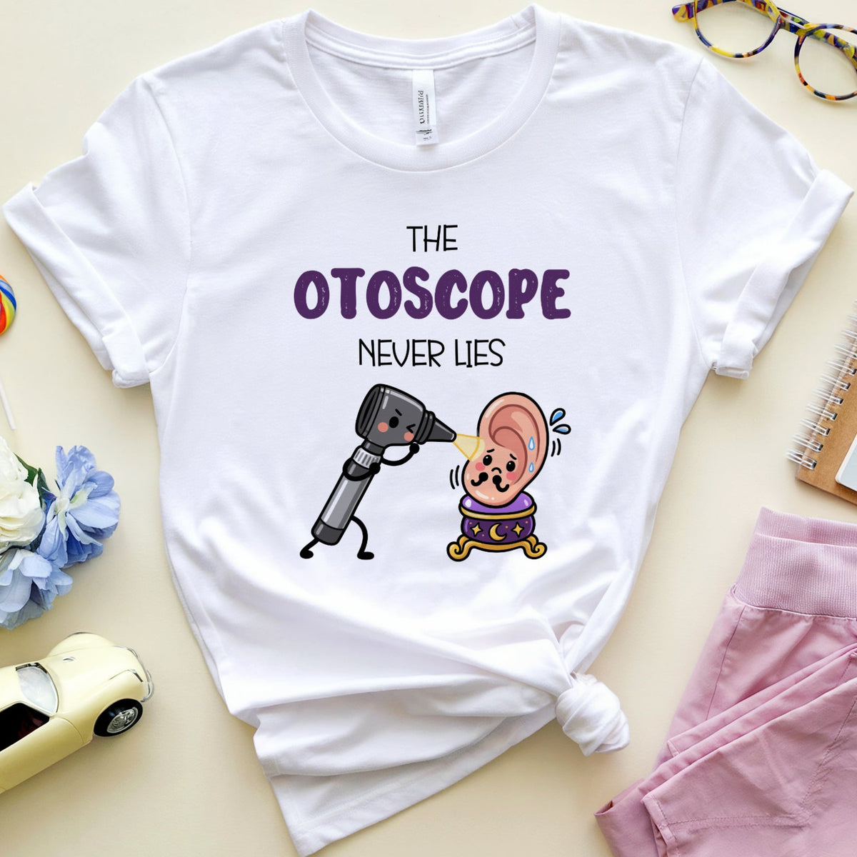 A white graphic t-shirt laid flat on a light beige background, styled with pink pants, glasses, flowers, a notebook, a lollipop, and a small car. The shirt features the text 'THE OTOSCOPE NEVER LIES' above a cartoon character holding an otoscope.