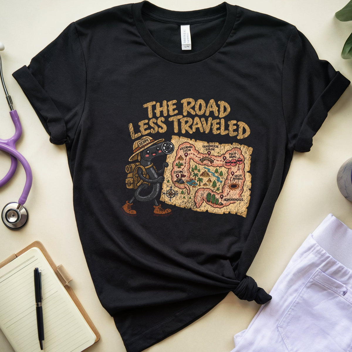 A black graphic t-shirt laid flat on a light beige surface, styled with a purple stethoscope, notebook, pen, folded white pants, a mug, and a plant. The t-shirt features a graphic of a dachshund wearing a cowboy hat next to a map, with the text 'THE ROAD LESS TRAVELED'.
