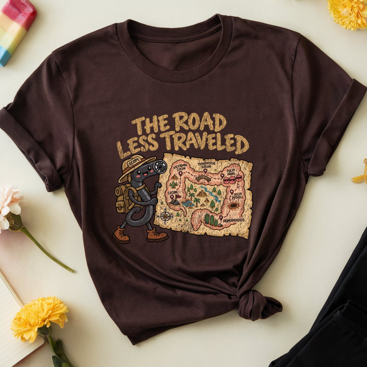 A dark brown graphic t-shirt with a knotted hem, featuring the text 'THE ROAD LESS TRAVELED' and an illustration of a map and a character. The t-shirt is styled in a flat lay on a light beige background with decorative props including flowers, a popsicle, glasses, and black pants.