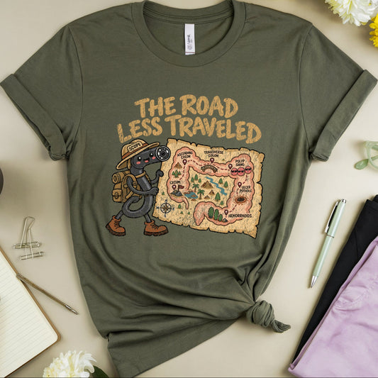 An olive green graphic t-shirt is styled in a flat lay on a beige background. The t-shirt features a cartoon graphic of a hiker next to a map with the text 'THE ROAD LESS TRAVELED'.
