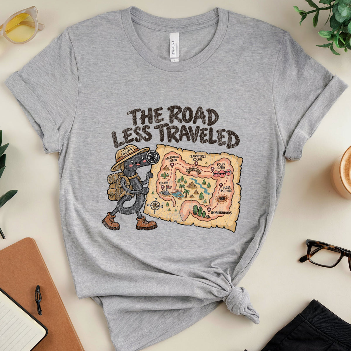 A heather gray graphic t-shirt with a knot at the hem, featuring a cartoon dinosaur and the text 'THE ROAD LESS TRAVELED' next to a map. The t-shirt is styled in a flat lay with sunglasses, a notebook, a coffee cup, and glasses on a light beige background.
