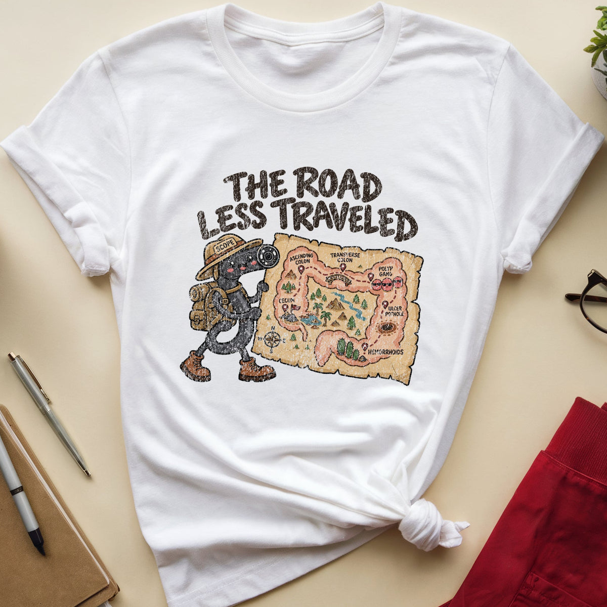 A white graphic t-shirt featuring the text 'THE ROAD LESS TRAVELED' and an illustration of a hiker with a map, styled in a flat lay with red pants, glasses, a notebook, and a plant on a beige background.