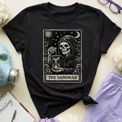 A black graphic T-shirt laid flat on a light beige surface, surrounded by decorative props including white and purple flowers, purple eyeglasses, a light blue gas mask, a notebook, and lavender scrubs. The shirt features a large tarot-style 'The Sandman' illustration on the front.