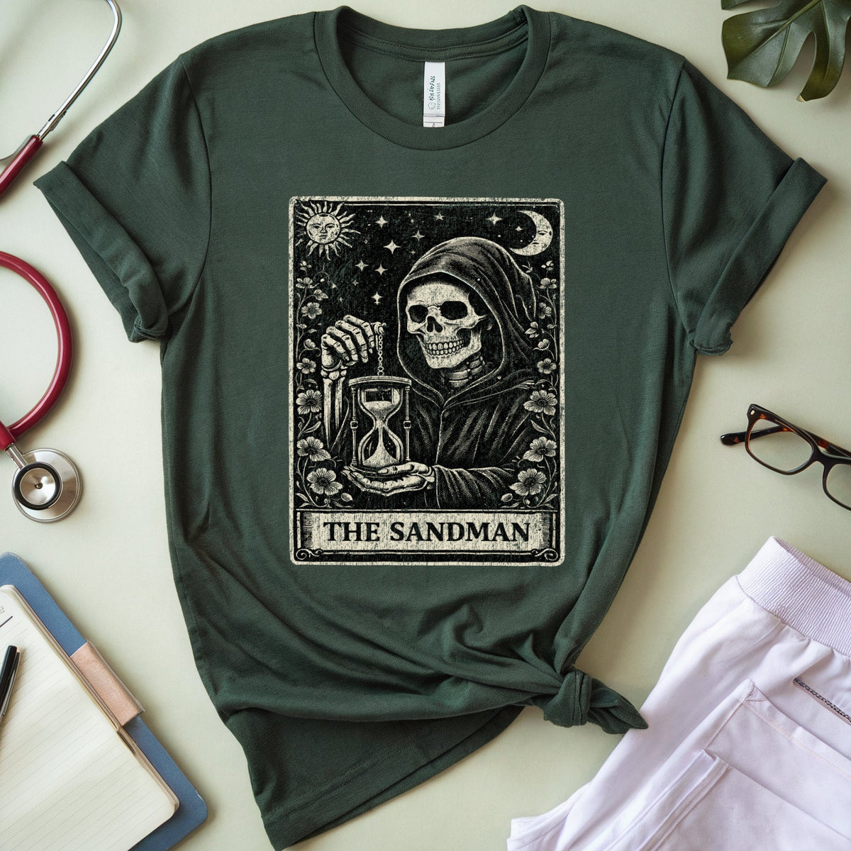 A dark forest green graphic t-shirt featuring 'The Sandman' tarot card design, styled in a flat-lay arrangement with a red stethoscope, notebook, glasses, and light purple pants on a light beige background.