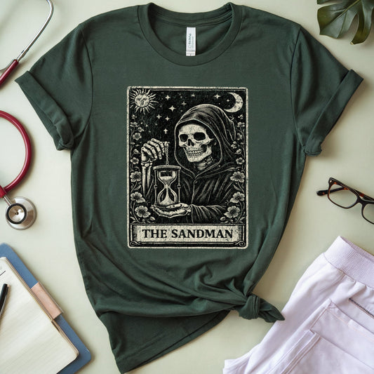 A dark forest green graphic t-shirt featuring 'The Sandman' tarot card design, styled in a flat-lay arrangement with a red stethoscope, notebook, glasses, and light purple pants on a light beige background.