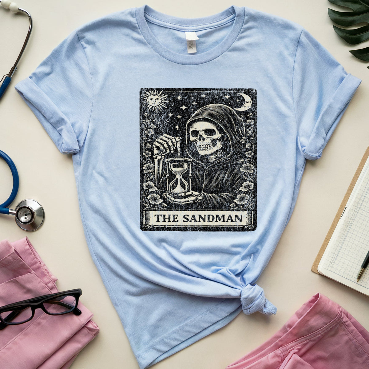 A light blue short-sleeve T-shirt laid flat on a beige surface, styled with pink scrubs, a stethoscope, eyeglasses, a notebook, and a green leaf. The shirt has a large black tarot-style graphic on the chest reading 'THE SANDMAN.
