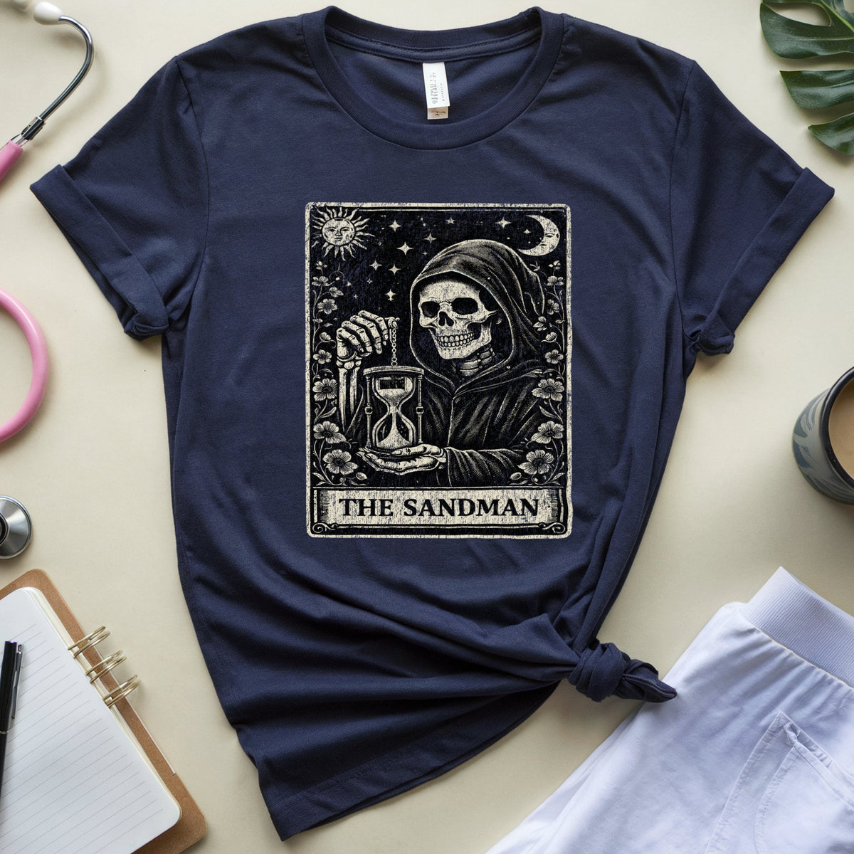 A navy blue graphic t-shirt featuring 'The Sandman' tarot card artwork, styled in a flat-lay arrangement with a pink stethoscope, notebook, plant leaf, and coffee cup on a beige background.