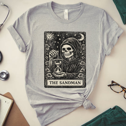 A heather gray short-sleeve T-shirt laid flat on a light beige surface, styled with a stethoscope, notebook, glasses, folded green fabric, and a knot at the hem. The shirt features a large black tarot-style graphic of The Sandman holding an hourglass under a crescent moon and stars, with the text 'THE SANDMAN' below.
