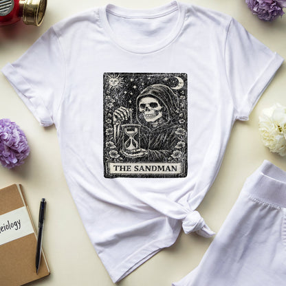 A white graphic T-shirt laid flat on a light cream background, styled with a red gas mask, blue glasses, flowers, a notebook, and white shorts. The shirt features a large black tarot-style 'The Sandman' illustration on the front.