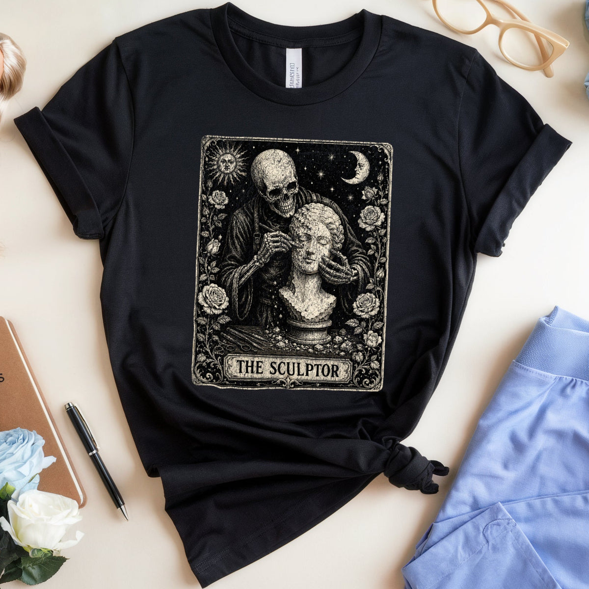 A black short-sleeve T-shirt laid flat on a light beige surface, featuring a large rectangular graphic print with a skeleton sculptor working on a female bust under a crescent moon, labeled 'THE SCULPTOR.' The shirt is styled in a flat lay with blue roses, glasses, a doll, a notebook, a pen, and light blue pants nearby.