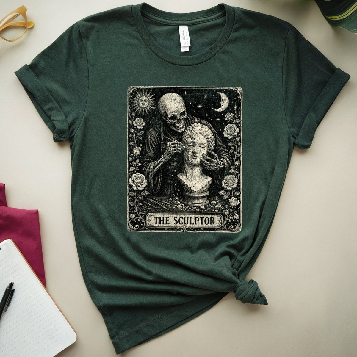 A dark green graphic t-shirt featuring a tarot card style illustration of a skeleton sculptor working on a female bust, labeled 'THE SCULPTOR'. The t-shirt is styled in a flat-lay arrangement on a light beige background with decorative props including a notebook, glasses, leaves, and a magenta cloth.