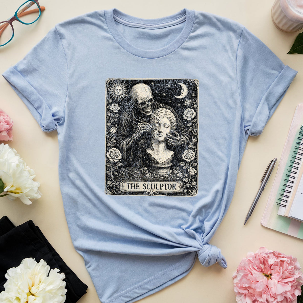 A light blue short-sleeve T-shirt laid flat on a beige surface, styled with flowers, glasses, a notebook, a pen, and a cup. The shirt has a large black-and-white tarot-style graphic on the chest reading “THE SCULPTOR,” and the lower hem is tied in a knot.