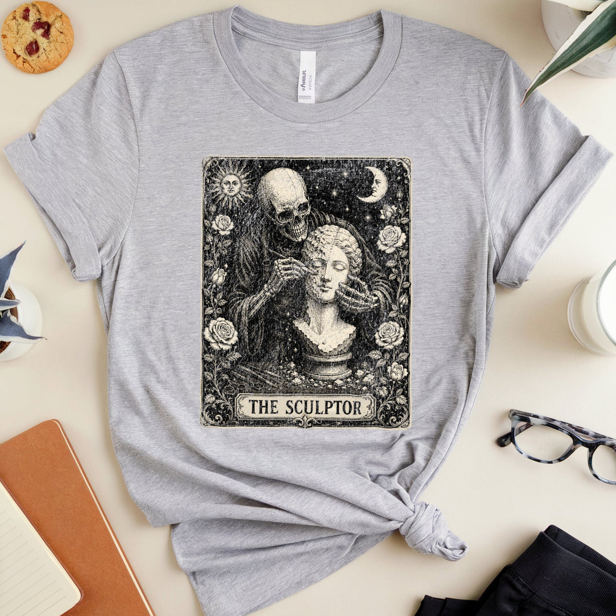 A heather gray short-sleeve T-shirt with a black-and-white gothic graphic of a sculptor and statue, styled in a flat lay with notebooks, glasses, a plant, and a candle on a light beige background.