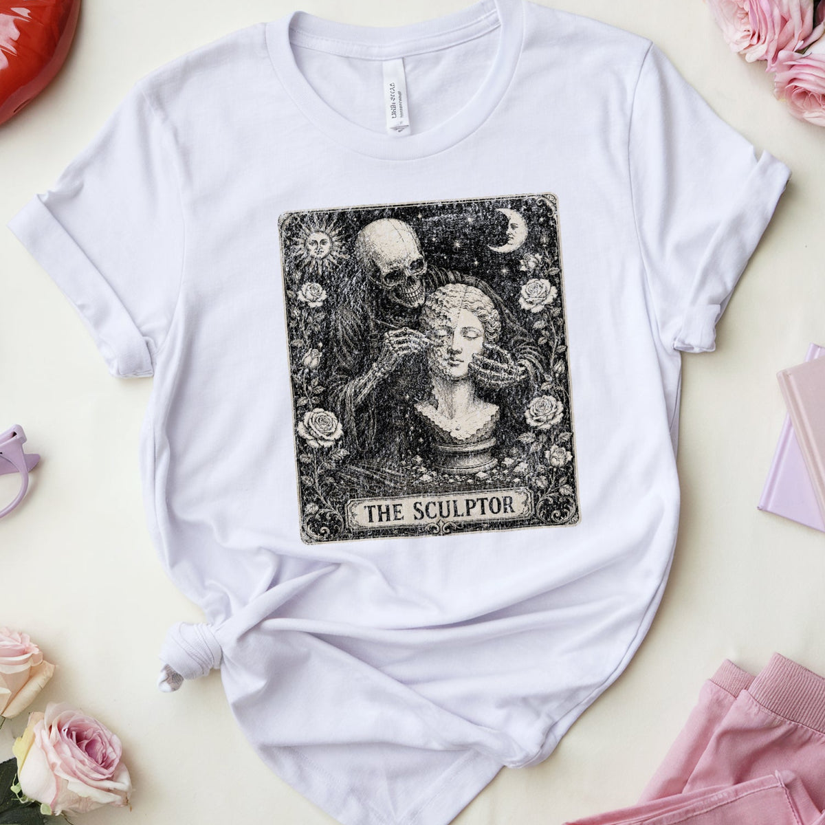 A white graphic t-shirt featuring a black and white 'The Sculptor' tarot card design, styled in a flat lay with pink accessories, glasses, lipstick, and roses on a light cream background.