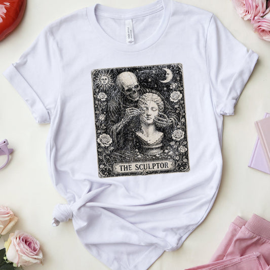 A white graphic t-shirt featuring a black and white 'The Sculptor' tarot card design, styled in a flat lay with pink accessories, glasses, lipstick, and roses on a light cream background.