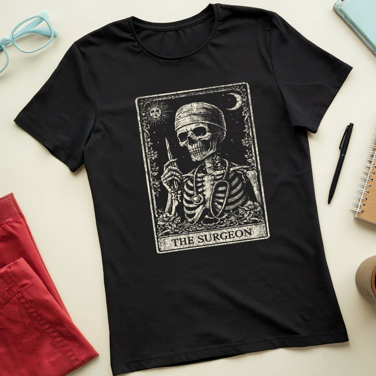 A black short-sleeve t-shirt with a vintage-style tarot card graphic of a skeleton labeled 'THE SURGEON'. The shirt is presented in a styled flat-lay arrangement on a light beige background, surrounded by props including red shorts, light blue glasses, a notebook, a pen, and a coffee cup.