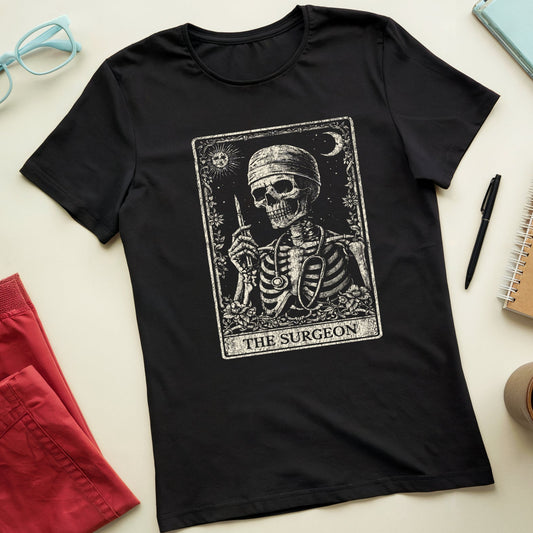 A black short-sleeve t-shirt with a vintage-style tarot card graphic of a skeleton labeled 'THE SURGEON'. The shirt is presented in a styled flat-lay arrangement on a light beige background, surrounded by props including red shorts, light blue glasses, a notebook, a pen, and a coffee cup.