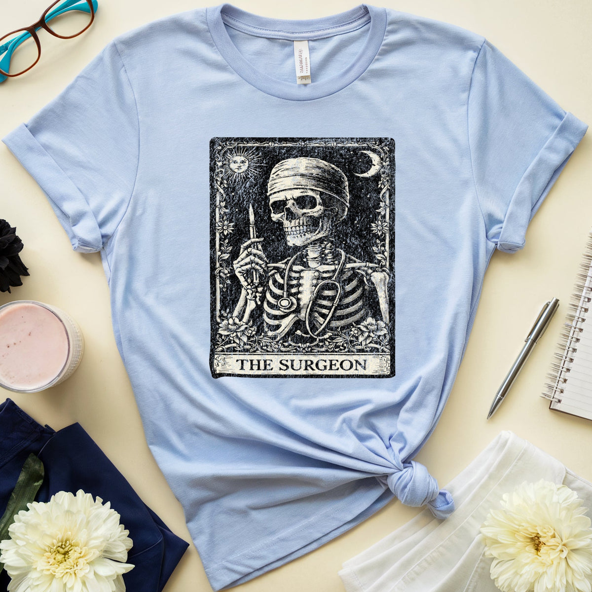 A light blue short-sleeve T-shirt laid flat on a cream background, styled with glasses, a notebook, flowers, a candle, and navy shorts. The shirt has a large black tarot-style graphic of a skeleton labeled 'THE SURGEON' on the chest.