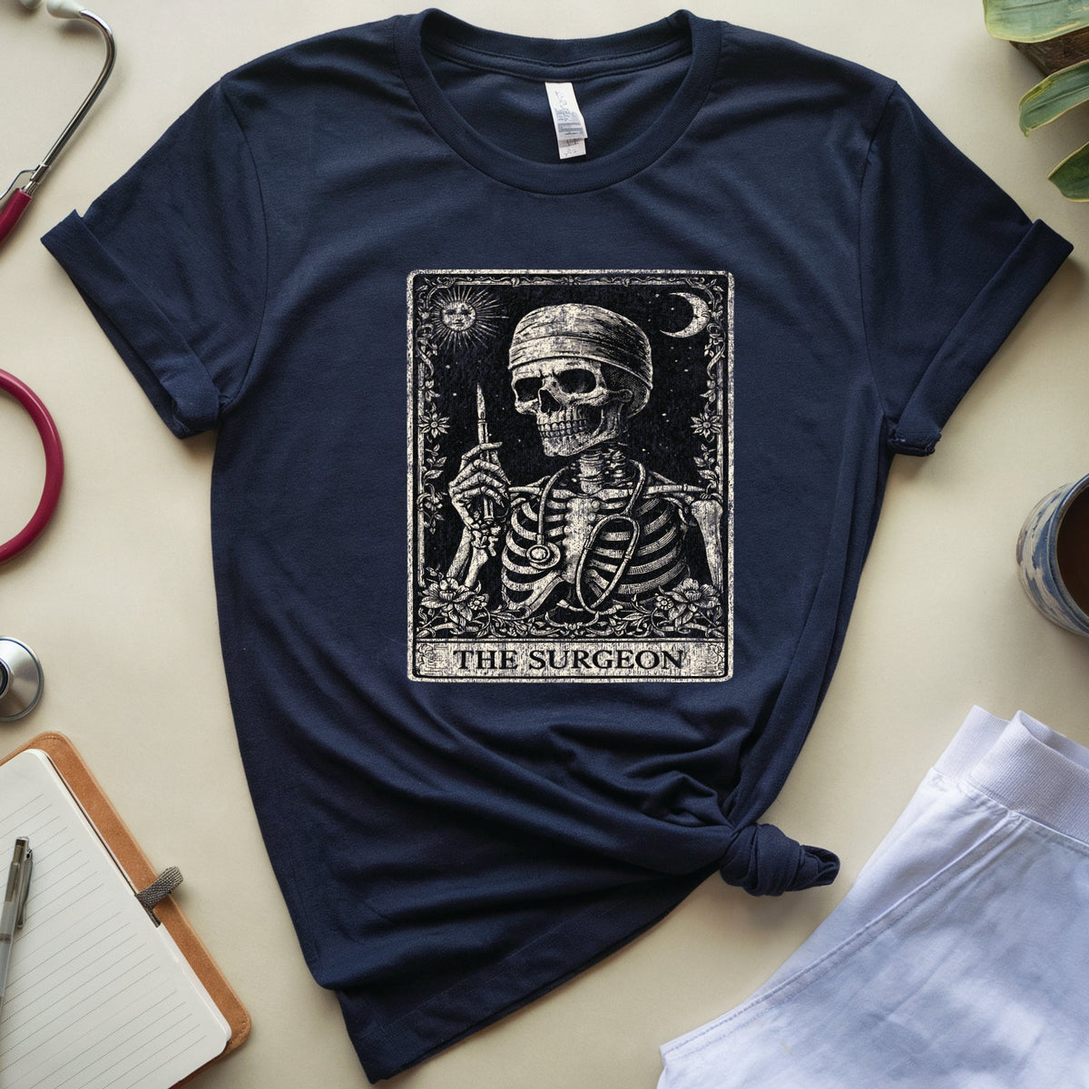A navy blue graphic t-shirt featuring a tarot card design of 'The Surgeon' with a skeleton holding a scalpel. The t-shirt is styled in a flat-lay arrangement on a beige surface with a stethoscope, notebook, plant leaves, and a cup of coffee.