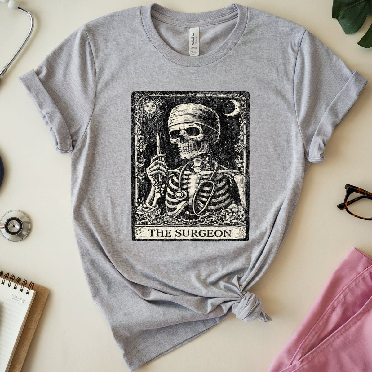 A heather gray short-sleeve T-shirt with a black tarot-style graphic of a skeleton labeled 'THE SURGEON,' styled in a flat lay with medical props including a stethoscope, notebook, pen, glasses, and pink scrubs on a light beige background.