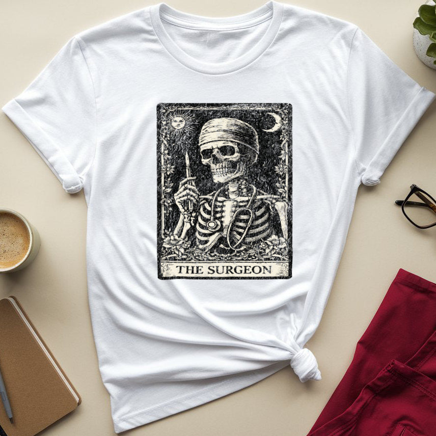A white short-sleeve T-shirt laid flat on a beige surface, styled with a black tarot-style graphic of a skeleton labeled 'THE SURGEON' on the chest. Surrounding props include a cup of coffee, a notebook with a pen, eyeglasses, a folded red garment, and a small potted succulent.