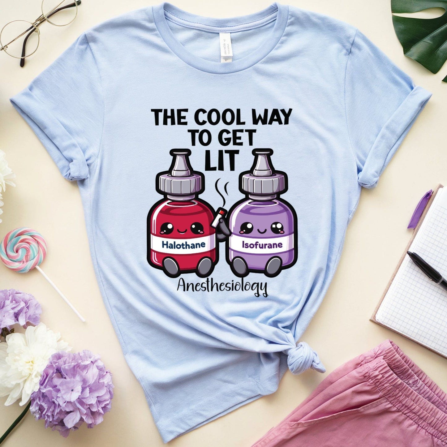 Light blue t-shirt with cartoon anesthesia bottles and text on a light surface with flowers and glasses.