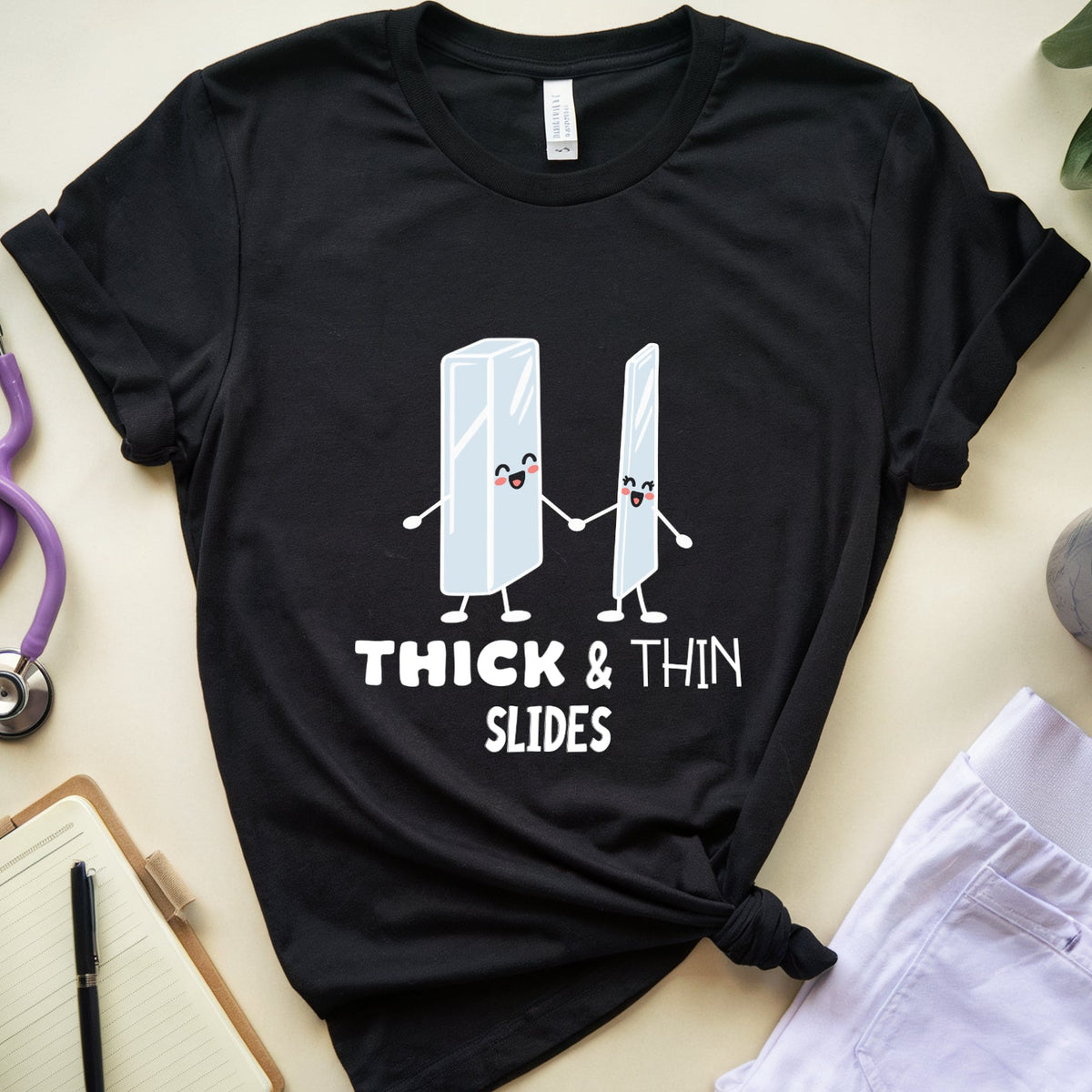 A black graphic t-shirt featuring two cartooning pencils holding hands with the text 'THICK & THIN SLIDES' underneath. The t-shirt is styled in a flat-lay arrangement on a light beige background, surrounded by a purple stethoscope, a notebook, a pen, a cup of coffee, and a white lab coat.