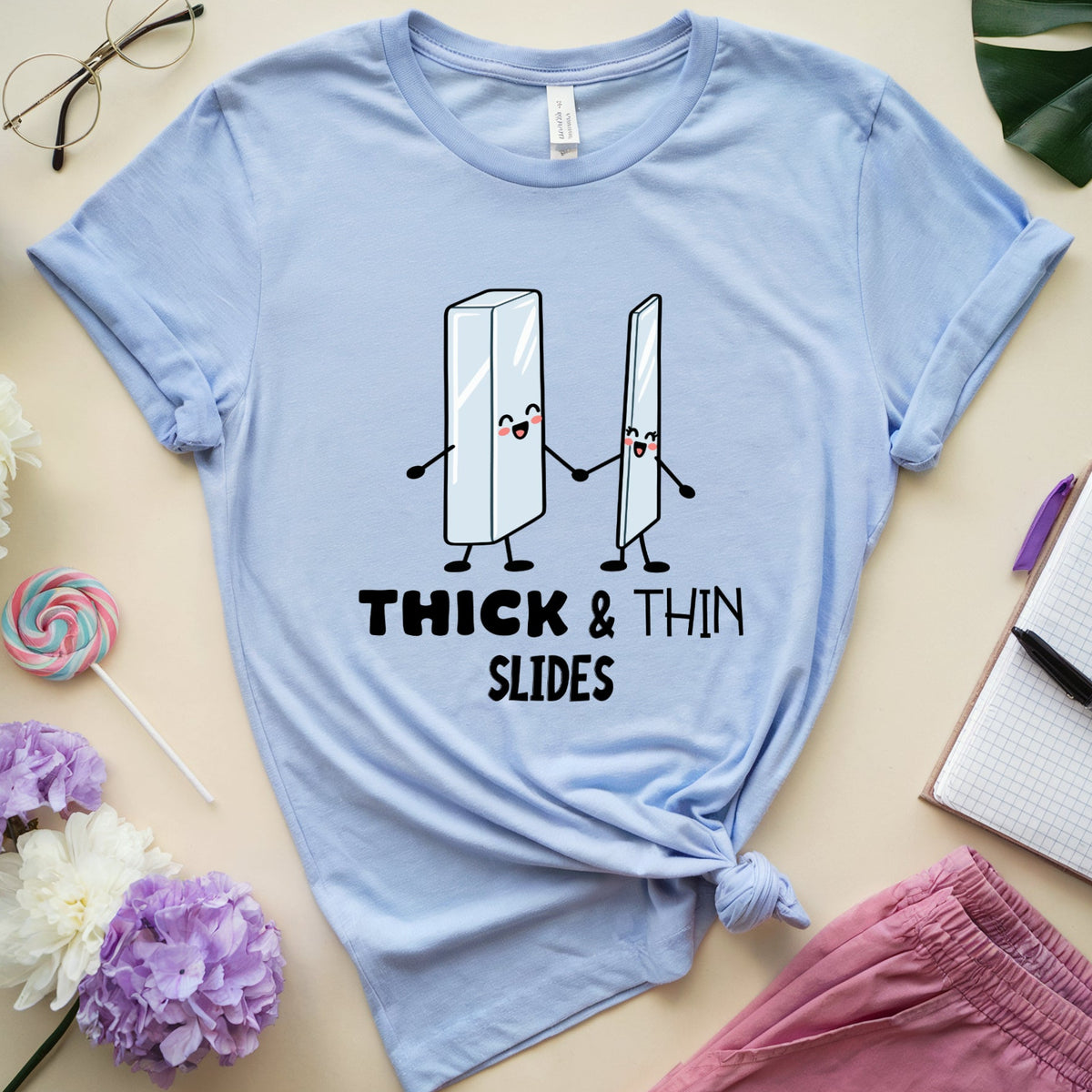 A light blue graphic t-shirt laid flat on a beige surface, surrounded by decorative props including flowers, glasses, a notebook, a lollipop, and pink pants. The shirt features a cartoon graphic of two sliding blocks holding hands with the text 'THICK & THIN SLIDES'.
