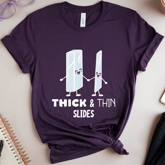 A dark purple graphic t-shirt featuring cartoon ice cream scoops and the text 'THICK & THIN SLIDES' in white. The t-shirt is styled in a flat-lay arrangement on a beige background with a pair of black shorts, a cup of latte, purple glasses, and notebooks.