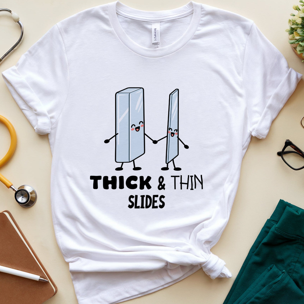 A white graphic t-shirt featuring two cartoon slides holding hands with the text 'THICK & THIN SLIDES' underneath. The t-shirt is styled in a flat-lay arrangement on a beige background, surrounded by medical-themed props including a stethoscope, notebook, glasses, and teal scrubs.