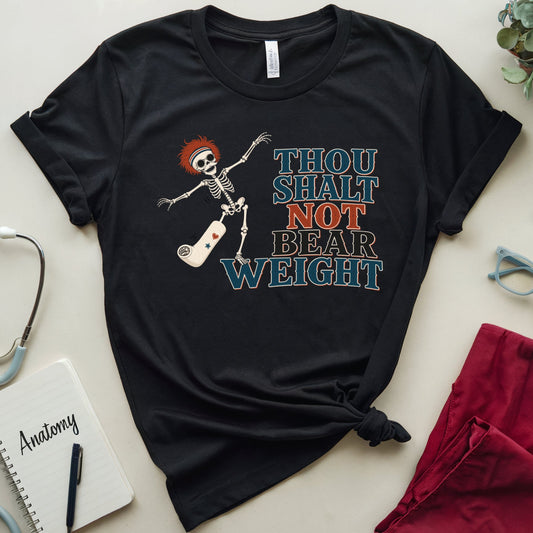 A black graphic t-shirt featuring a skeleton and the text 'THOU SHALT NOT BEAR WEIGHT' in blue and orange, styled in a flat lay with medical-themed props including a stethoscope, notebook, glasses, and red scrubs on a light beige background.